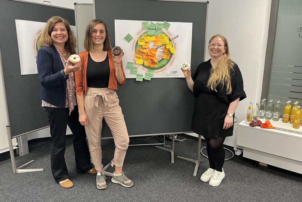 Gruppenfoto mit Frau Dr. Annekatrin Meißner, Nelly Rahimy und Sina Kehrwieder, jeweils mit einem Donut in der Hand. Im Hintergrund eine Pinnwand mit Ideen zu ökologischen und sozialen Projekten für Passau.