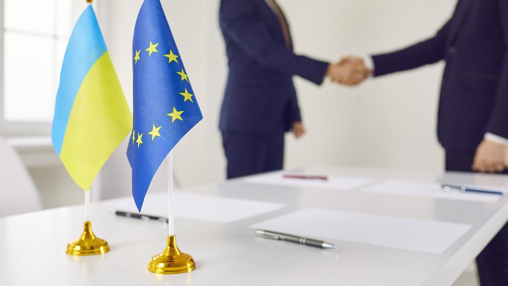 Ukrainische und EU-Flagge vor Politikern die sich die Hände schütteln.