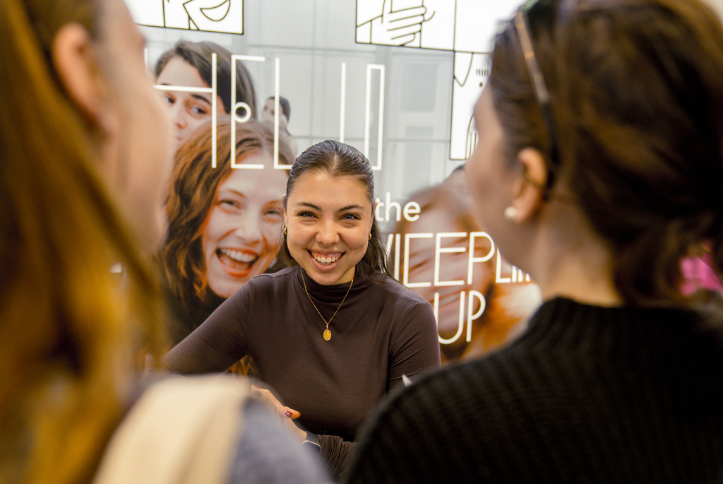 Auf diesem Bild sieht man eine Ausstellerin auf der Campus meets Company 2024 die lächelt. Im Vordergrund unscharf zu sehen sind zwei Hinterköpfe von Personen, die sich mit der Ausstellerin unterhalten.