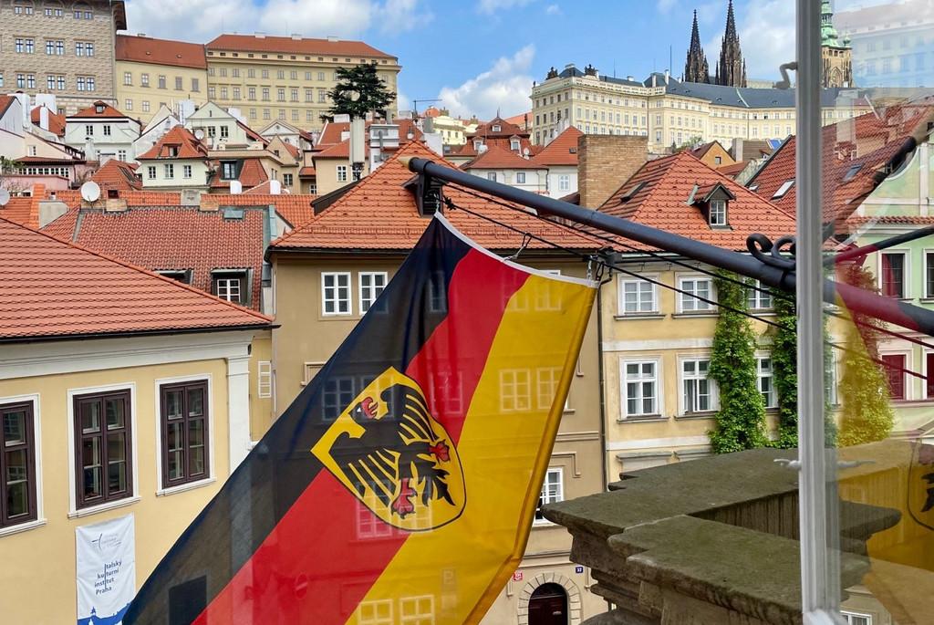 Auf diesem Bild sieht man eine deutsche Dienstflagge samt Adler, die aus der deutschen Botschaft in Prag nach draußen weht.  Die Flagge ist an einem Metallmast befestigt, der schräg aus der Wand eines Gebäudes ragt. Das Bild wurde aus einem Fenster des Gebäudes aufgenommen. Im Hintergrund sieht man eine malerische Straße der Prager Altstadt, die mit Kopfsteinpflaster bedeckt ist. Links unten im Bild stehen zwei Menschen, die nach oben schauen und möglicherweise ein Foto machen. In der Nähe sind mehrere Autos geparkt. Im Hintergrund sieht man eine Reihe von historischen Gebäuden mit roten Ziegeldächern, die sich treppenartig den Hügel hinaufziehen. Einige dieser Gebäude sind hellgelb oder beige gestrichen. Am Horizont, hinter diesen Gebäuden, ragen die spitzen Türme der Prager Burg in den Himmel. Der Himmel ist klar und blau, mit einigen weißen Wolken.