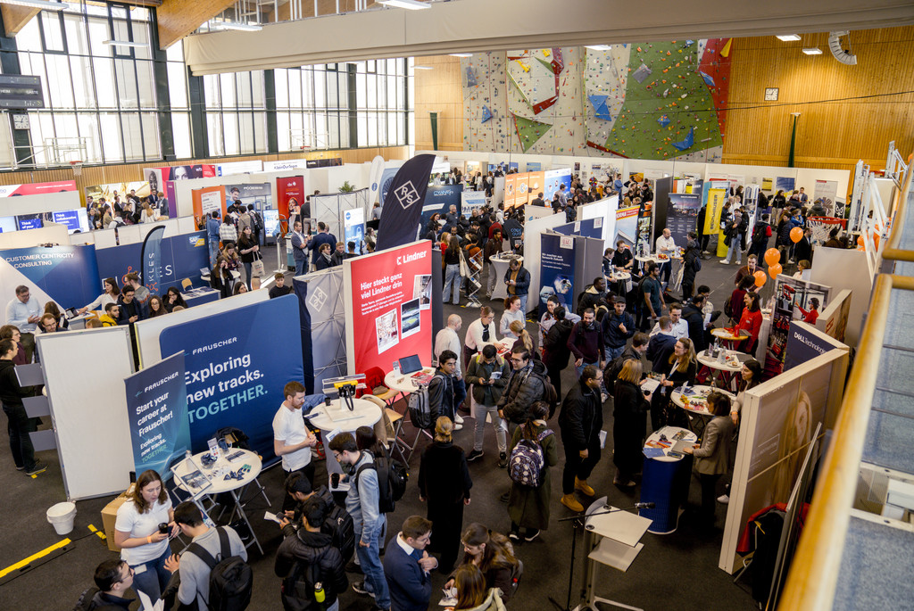 Auf dem Bild sieht man die Sporthalle der Universität Passau aus einer erhöhten Perspektive. Die Halle ist voller Menschen und Stände der einzelnen Unternehmen, die bei der Campus meets Company 2024 ausgestellt haben.