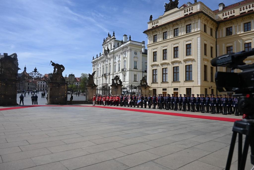 Staatsbesuch der deutschen Bundespräsidenten Frank-Walter Steinmeier in Prag. Zu sehen sind ein eingezäunter Vorplatz auf dem ein roter Teppich ausgerollt ist. Auf den Mauern sind unterschiedliche Figuren zu sehen. Rechts stehen Soldaten Spalier. Im Hintergrund sind zwei Häuser im Barock-Stil in weiß und beige zu sehen. Ganz rechts im Bild sieht man eine Kamera, die auf die Szenerie gerichtet ist.