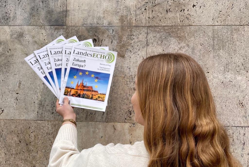 Auf dem Bild ist eine junge Frau von hinten zu sehen, die in der rechten Hand mehrere Zeitschriften hält. Die Person hat mittellanges, welliges, rötlich-blondes Haar, das bis zu den Schultern reicht. Sie trägt einen hellen, beige-weißen Pullover und steht vor einer grauen, marmorierten Steinwand. Die Zeitschriften, die die Frau in der Hand hält, sind identisch. Auf dem Titelblatt der Zeitschrift steht der Titel „LandesECHO“ in großen Buchstaben am oberen Rand. Direkt darunter steht der Text „Zukunft Europa?“ Die Titelseite zeigt ein Bild eines großen, beleuchteten Gebäudes, das im Dämmerlicht aufgenommen wurde, vor einem Himmel, der von mehreren gelben Sternen übersät ist – möglicherweise eine Anspielung auf die Europaflagge.