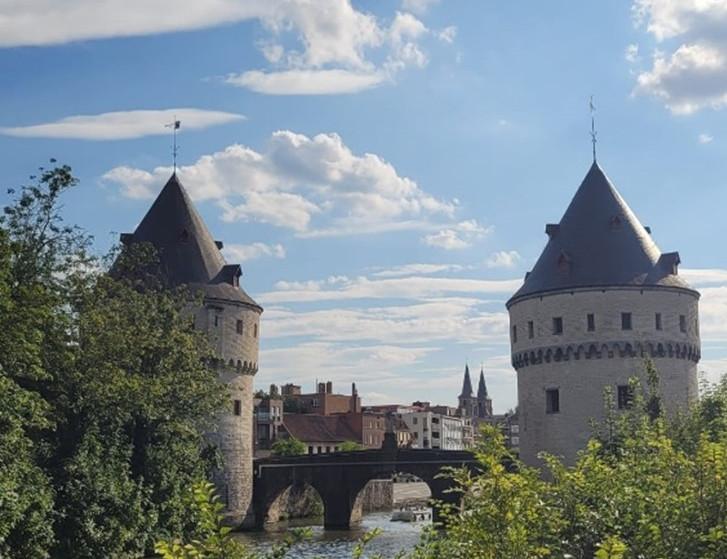 Das Bild zeigt die historischen Broeltürme in Kortrijk, Belgien. Zwei runde, mittelalterliche Türme mit kegelförmigen Dächern stehen am Ufer eines Flusses und sind durch eine Steinbrücke verbunden. Die Szene wird von Bäumen und Büschen im Vordergrund umrahmt, und der Himmel ist mit einigen Wolken bedeckt, was eine friedliche Atmosphäre schafft.