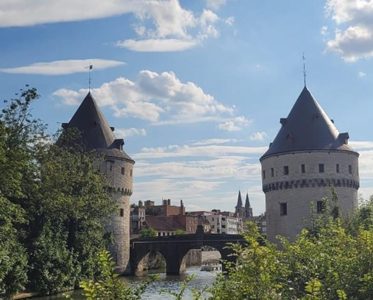 Das Bild zeigt die historischen Broeltürme in Kortrijk, Belgien. Zwei runde, mittelalterliche Türme mit kegelförmigen Dächern stehen am Ufer eines Flusses und sind durch eine Steinbrücke verbunden. Die Szene wird von Bäumen und Büschen im Vordergrund umrahmt, und der Himmel ist mit einigen Wolken bedeckt, was eine friedliche Atmosphäre schafft.