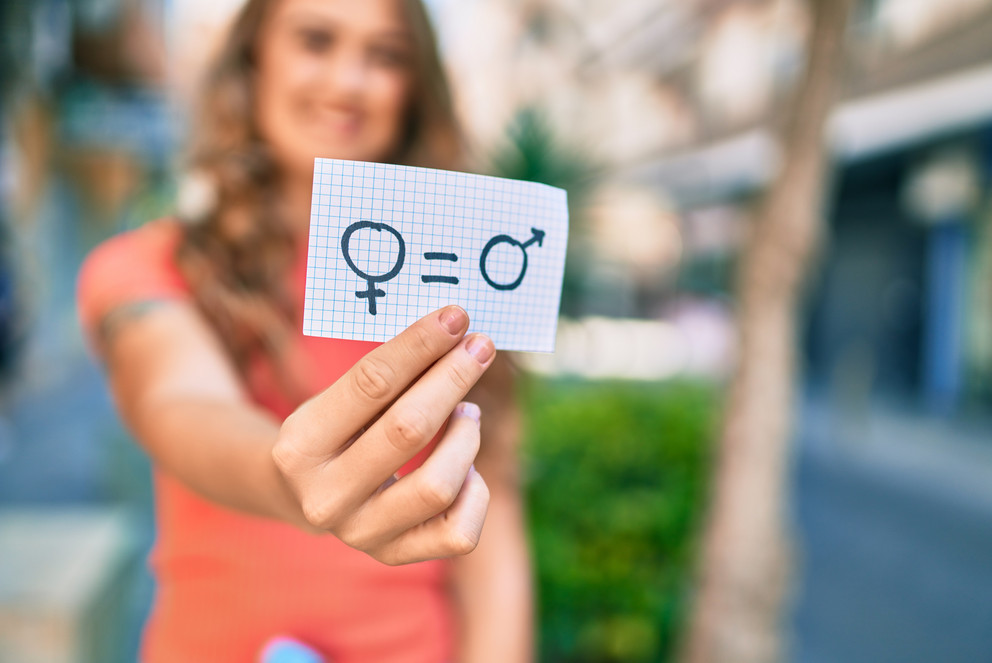 Eine Frau, die man nur verschwommen im Hintergrund erkennen kann, hält einen Zettel mit einem Symbol für Geschlechtergleichheit in die Kamera.