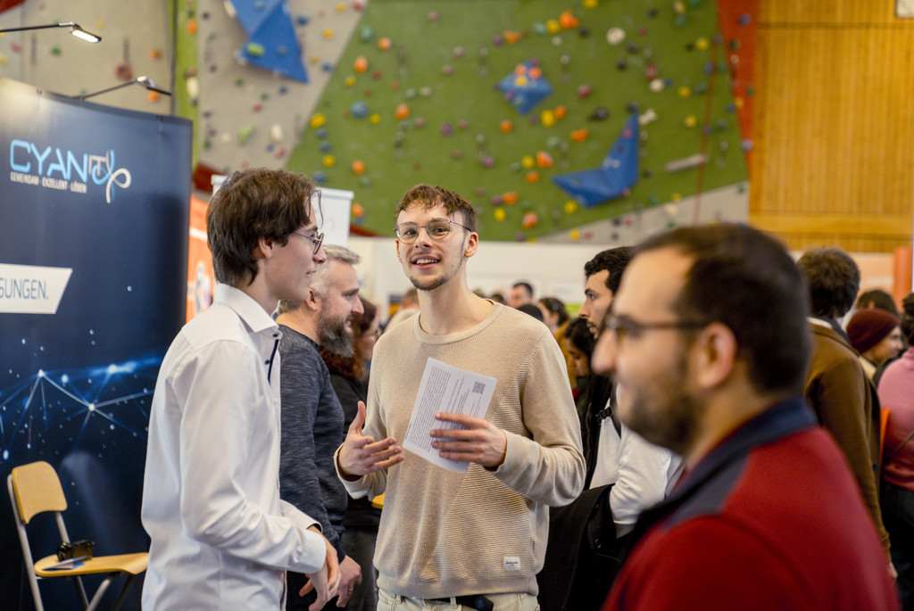 Im Vordergrund sieht man unscharf einen Mann. Dahinter sieht man scharf einen Aussteller und einen Studenten, die sich gerade unterhalten. Im Hintergrund sieht man die Kletterwand der Sporthalle an der Univeristät Passau.